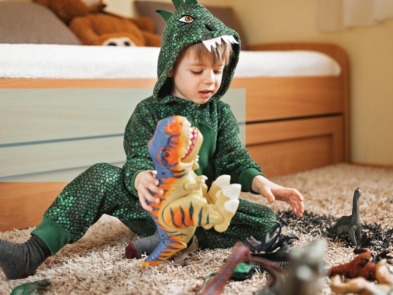 Barn i grøn dinosaurdragt leger med dinosaurfigurer på gulvet.
