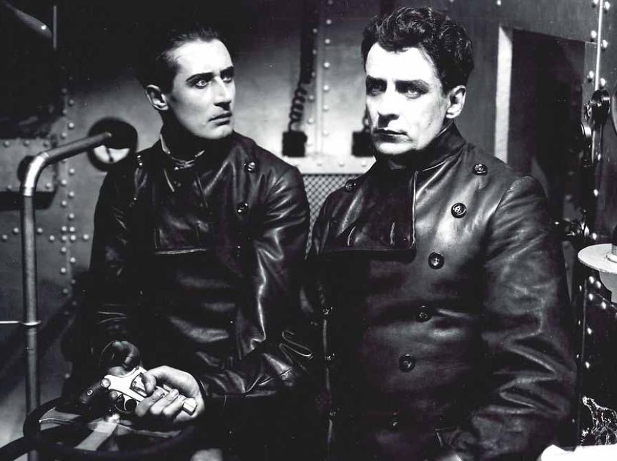 sort hvid billede fra stumfilmen Himmelskibet, to mænd i læderjakker sidder i et skib, stirrende intenst